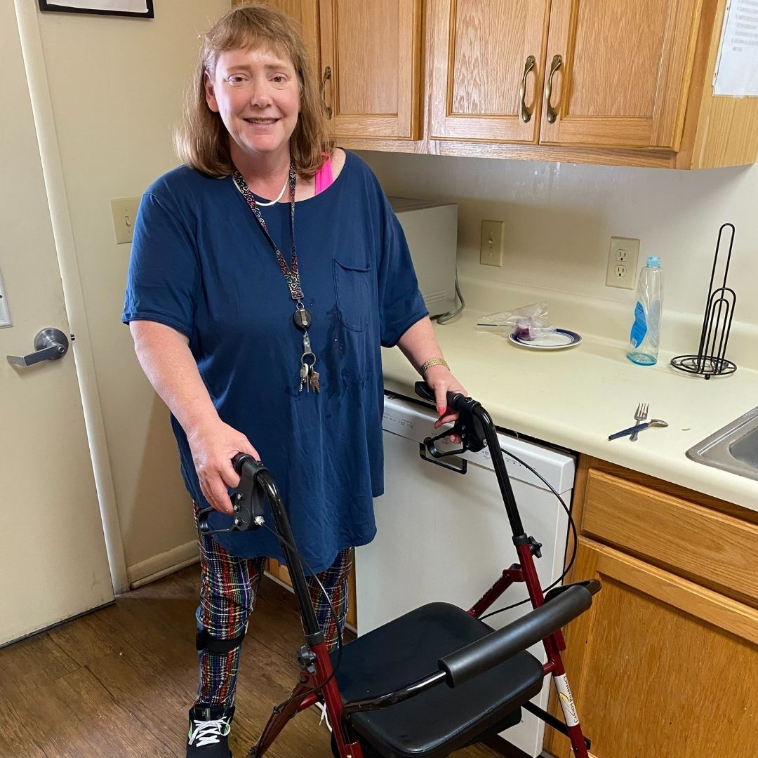 Windermere resident Stephanie Williams using her rolling walker wearing a blue top and printed pants in the home's kitchen.